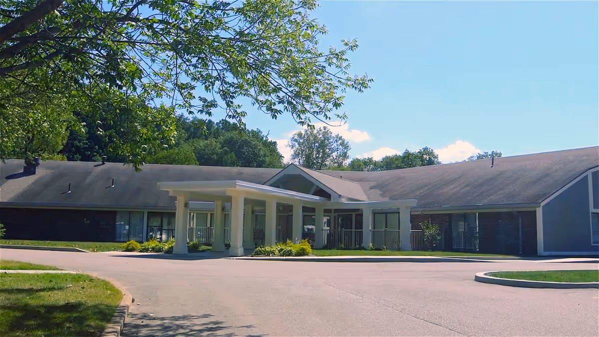Front entrance of a single-story senior living building with a covered porte-cochère, driveway, and surrounding trees.