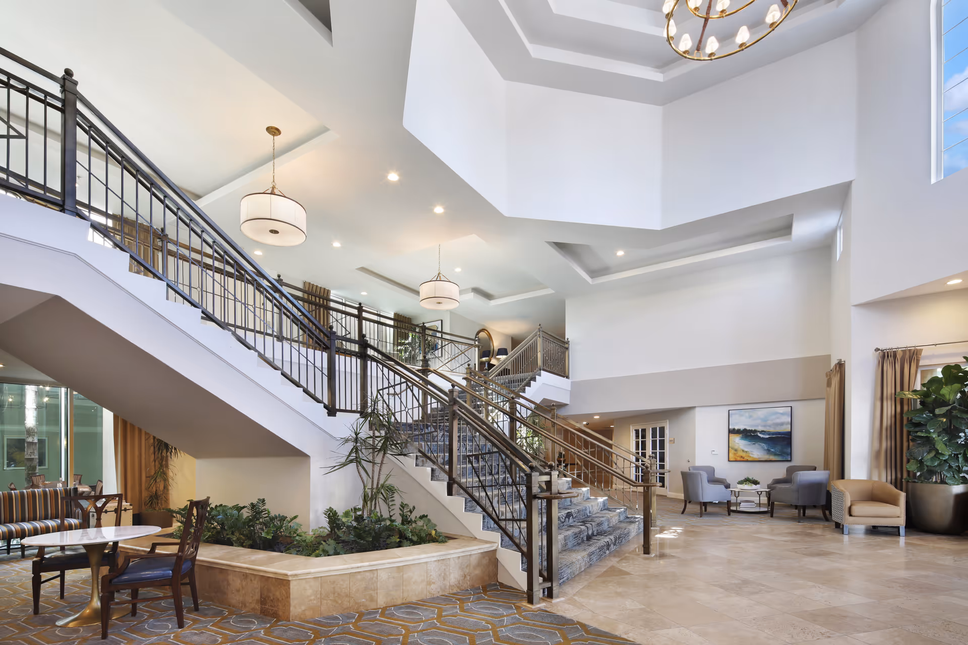 Spacious and well-lit interior of a senior living facility featuring a wide staircase with metal railings and carpeted steps, a seating area with chairs and a small table, large windows allowing natural light, potted plants, and modern ceiling light fixtures.