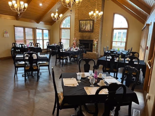 Dining room with multiple tables covered in black tablecloths, set with glasses, napkins, and utensils. The room features wooden floors, large windows with blinds, a brick fireplace with decorative bunny figures on the mantel, and chandeliers hanging from a vaulted wooden ceiling.