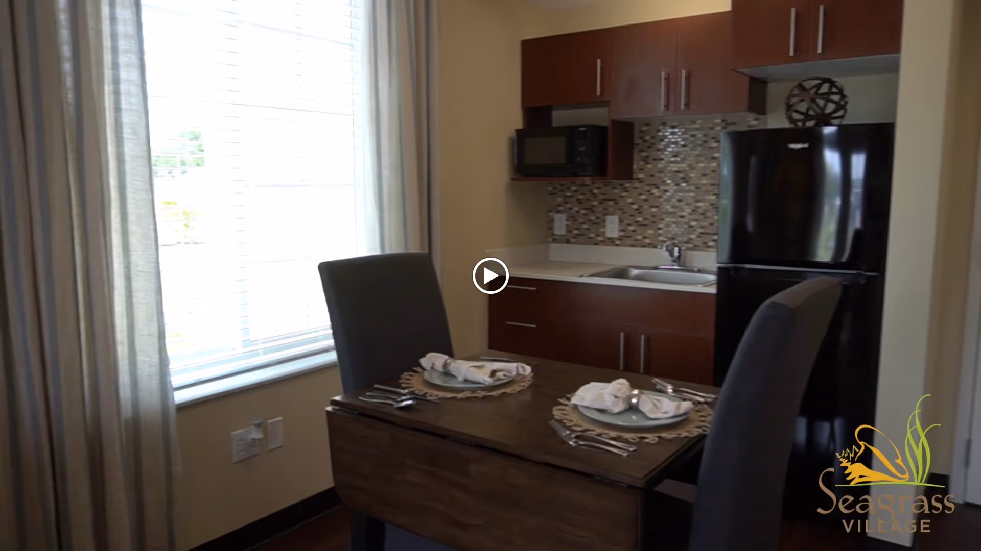A small kitchen and dining area with a wooden table set for two, featuring plates, napkins, and silverware. The kitchen has dark wood cabinets, a black refrigerator, a microwave, a sink, and a mosaic tile backsplash. A large window with blinds and curtains lets in natural light.