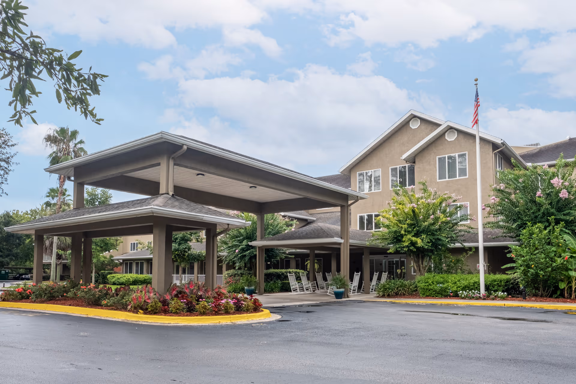 Exterior view of a senior living facility with a covered entrance, rocking chairs on the porch, landscaped flower beds, and an American flag on a flagpole under a partly cloudy sky.