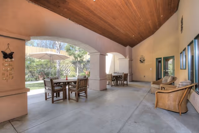 Covered outdoor patio area with wooden ceiling, featuring several wooden tables and chairs, wicker armchairs with cushions, and an umbrella providing shade over one table. The patio opens to a garden area with greenery and trees.