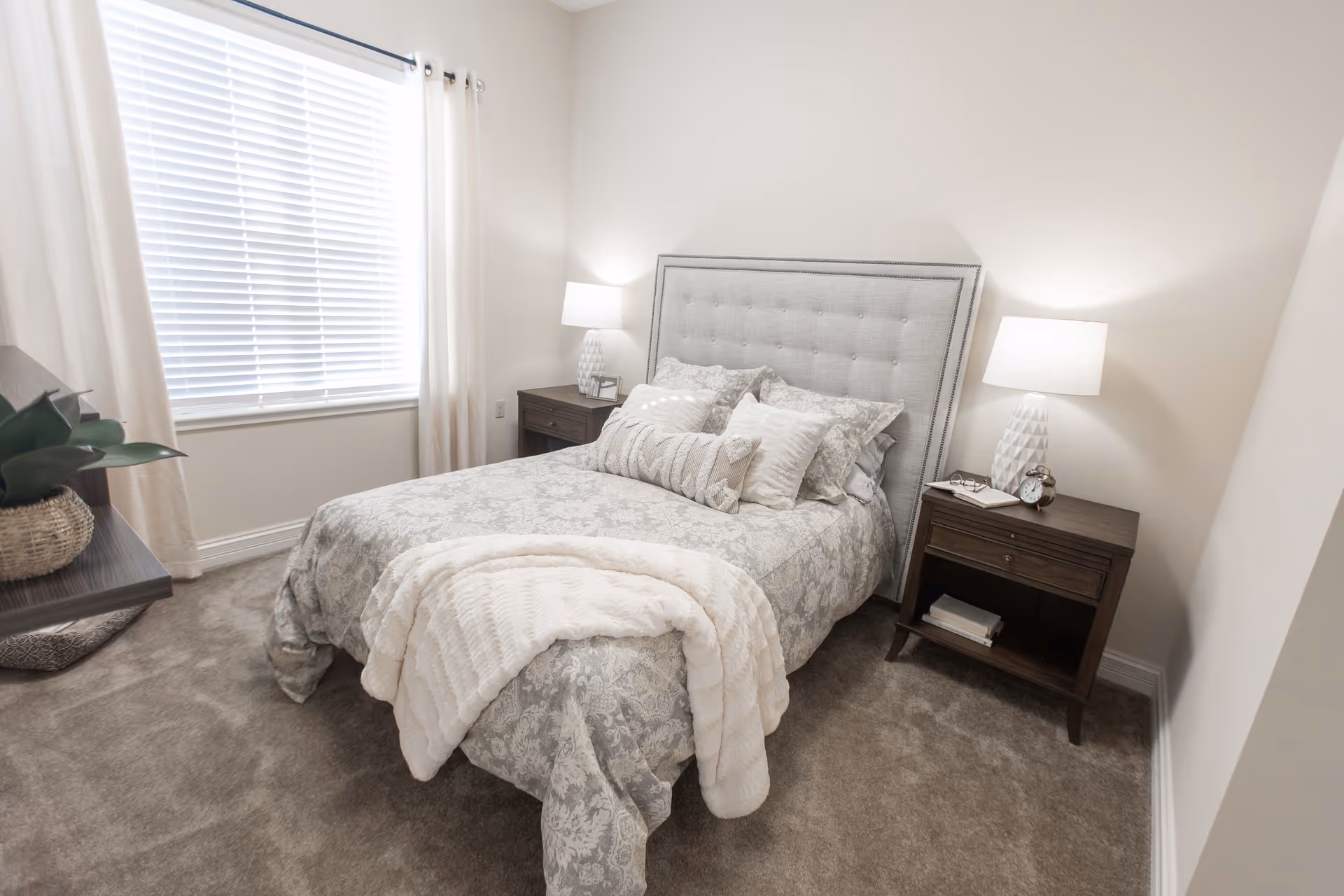 A cozy bedroom with a neatly made bed featuring a tufted headboard, multiple decorative pillows, and a soft throw blanket. There are two dark wooden nightstands on either side of the bed, each with a white table lamp. A window with white blinds and light curtains is on the left wall, and a small plant in a woven basket sits on a shelf near the window. The room has light-colored walls and carpeted flooring.