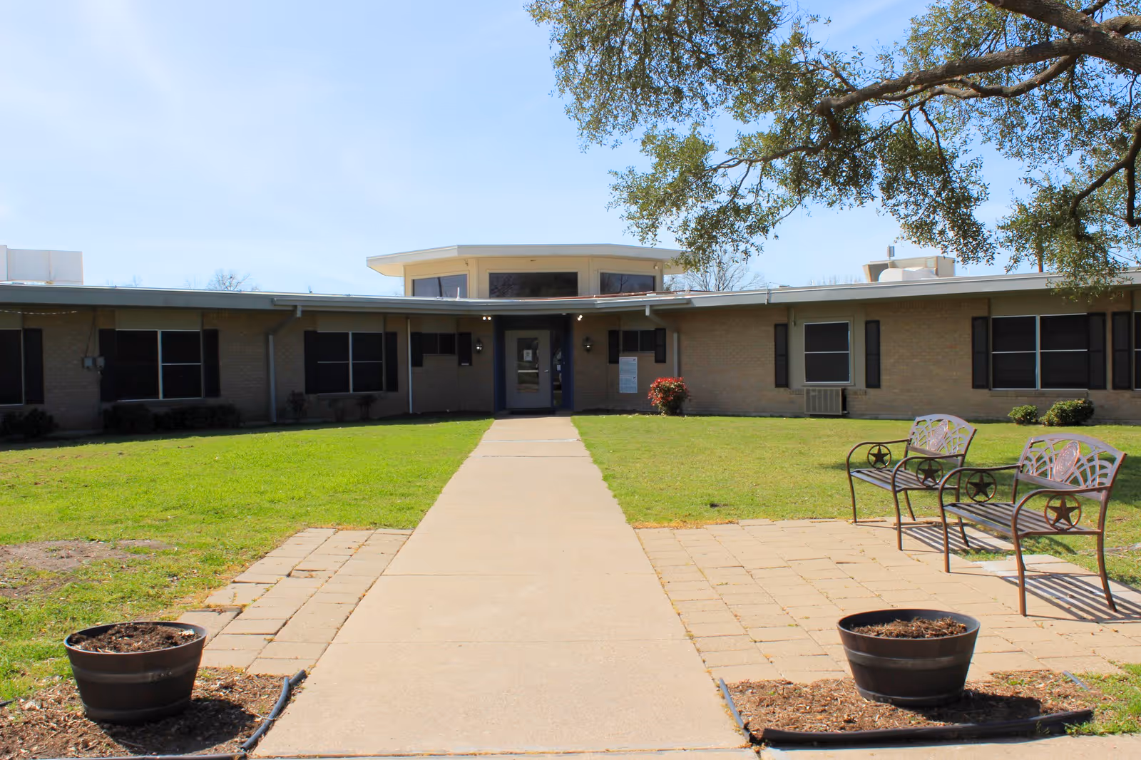 Front entrance of a single-story nursing facility with a paved walkway, lawn, benches and planters beneath a large tree.