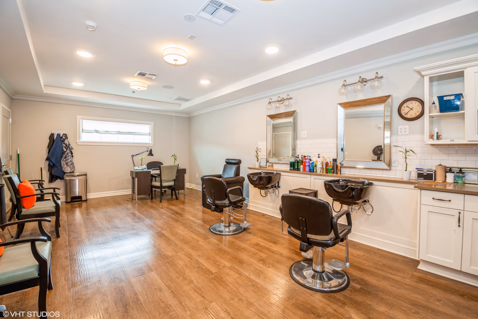 Interior view of a salon area in a senior living facility with two black salon chairs in front of two sinks and mirrors. The room has wooden flooring, white walls, and ceiling lights. There are chairs with orange cushions along one wall and a desk with two chairs near a window.