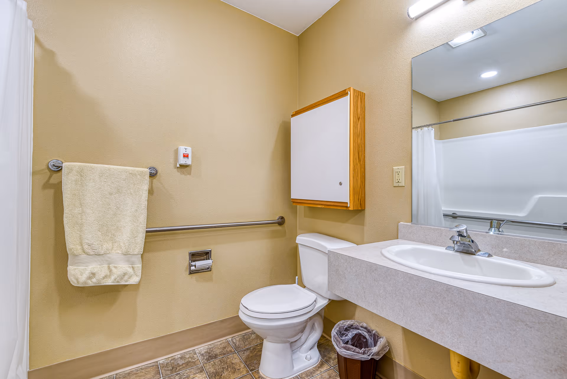 A clean bathroom with beige walls and tiled floor featuring a white toilet, a white sink with a countertop, a large mirror above the sink, a towel rack with a beige towel, a wall-mounted cabinet, a trash bin, and a shower area with a white curtain.