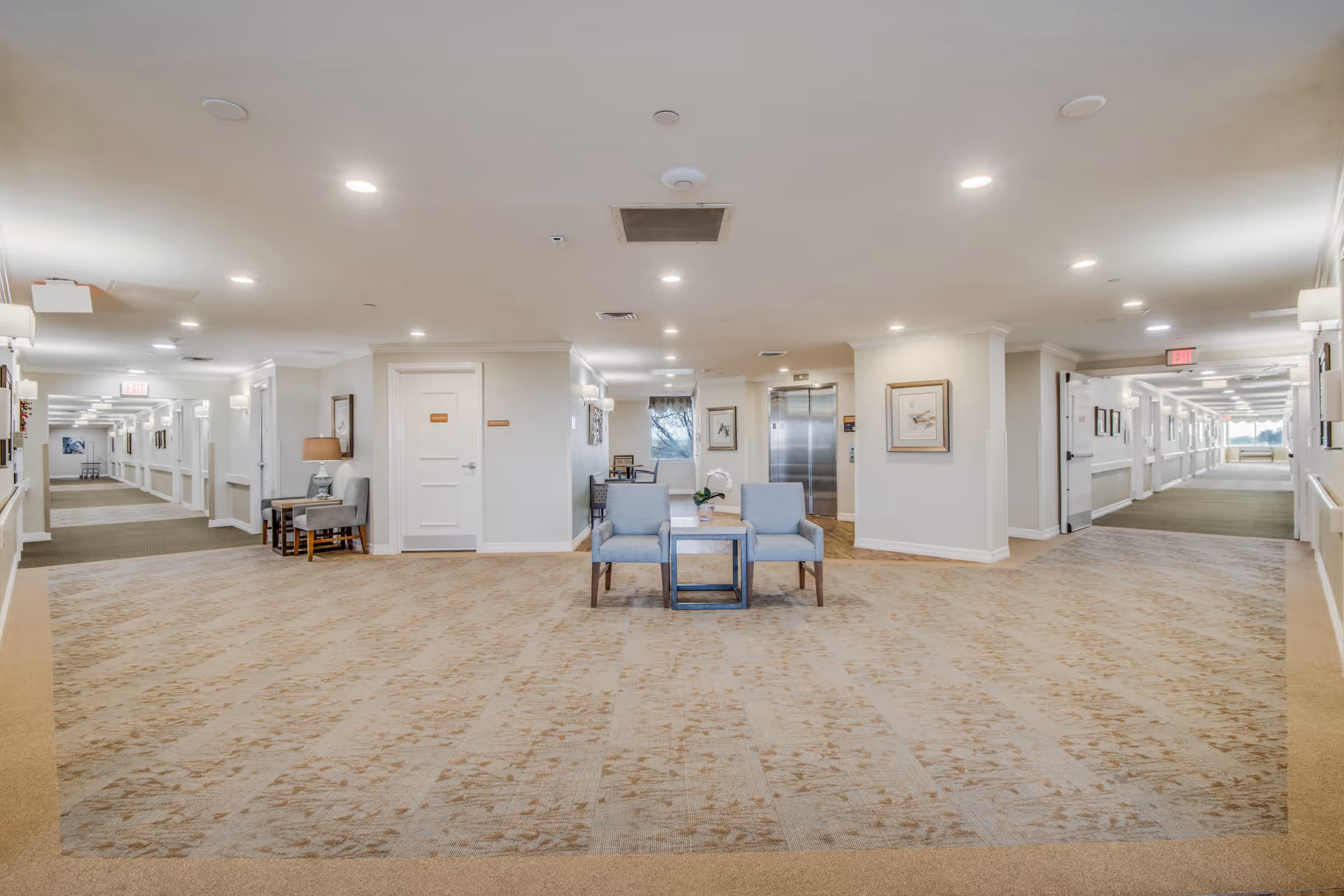 A spacious, well-lit hallway in a senior living facility with beige patterned carpet and white walls. Two blue armchairs with a small table between them are positioned in the center. The hallway extends to the left and right with multiple doors, framed artwork on the walls, and ceiling lights. An elevator is visible in the background, along with a window letting in natural light.
