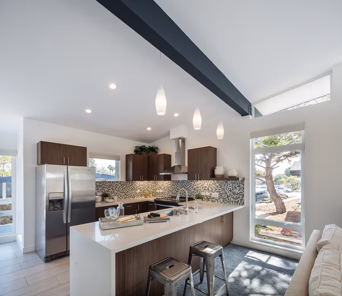 Modern kitchen with a large white island countertop, two metal stools, dark wood cabinets, stainless steel refrigerator and stove, mosaic tile backsplash, pendant lights hanging from the ceiling, and a large window showing outdoor trees and sunlight.