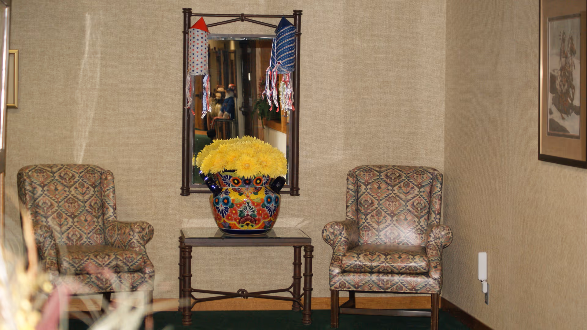 Two patterned armchairs flanking a small table topped with a colorful vase of yellow flowers and a mirror on a beige wall.