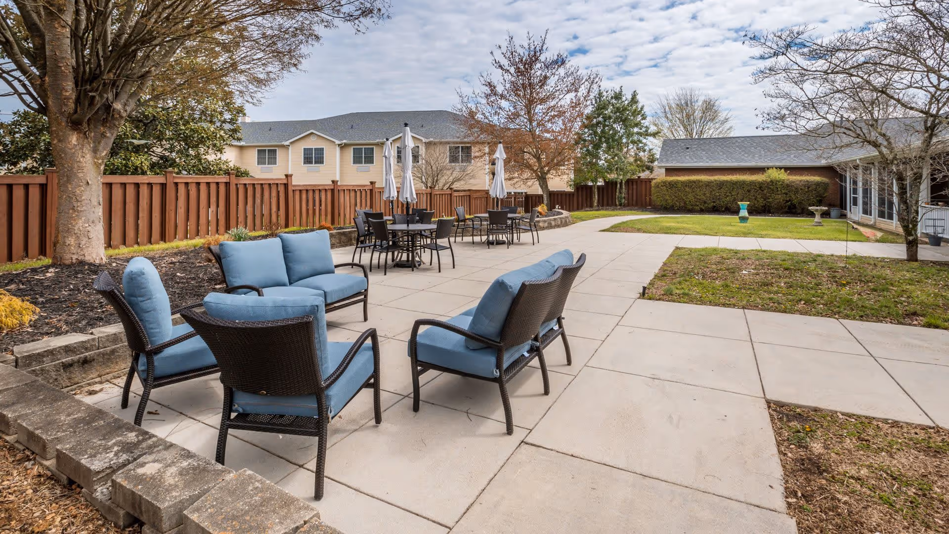 Outdoor patio area with several sets of black wicker chairs and sofas with blue cushions arranged around tables and open umbrellas. The patio is surrounded by a wooden fence, trees, and a grassy area with a few shrubs and a birdbath. Residential buildings are visible in the background under a partly cloudy sky.
