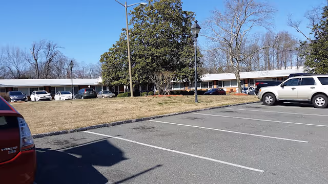 Parking lot and grassy lawn in front of a single-story senior living building with parked cars and trees under a clear blue sky.