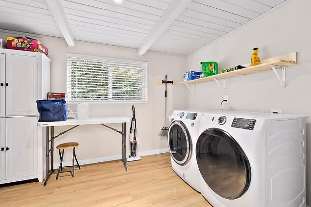 A bright laundry room with a front-loading washing machine and dryer side by side on the right. Above them is a wooden shelf holding laundry supplies. On the left side, there is a white folding table with a small stool underneath and a blue bag on top. A white cabinet stands next to the table. A window with white blinds lets in natural light, and cleaning tools like a broom and vacuum are placed against the wall.