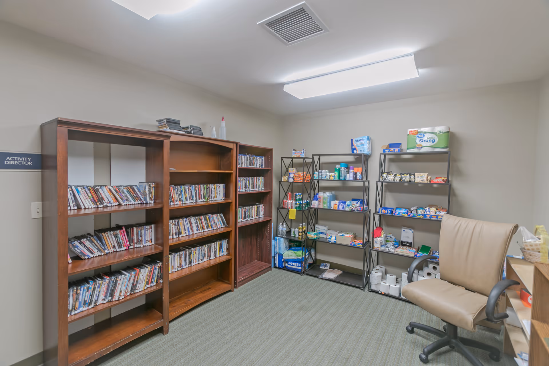 A small room with wooden shelves filled with DVDs on the left and metal shelves stocked with various supplies such as paper towels, snacks, and toiletries on the right. There is a beige office chair with armrests in the foreground and a sign on the wall that reads 'Activity Director'. The room has a carpeted floor and a ceiling light.