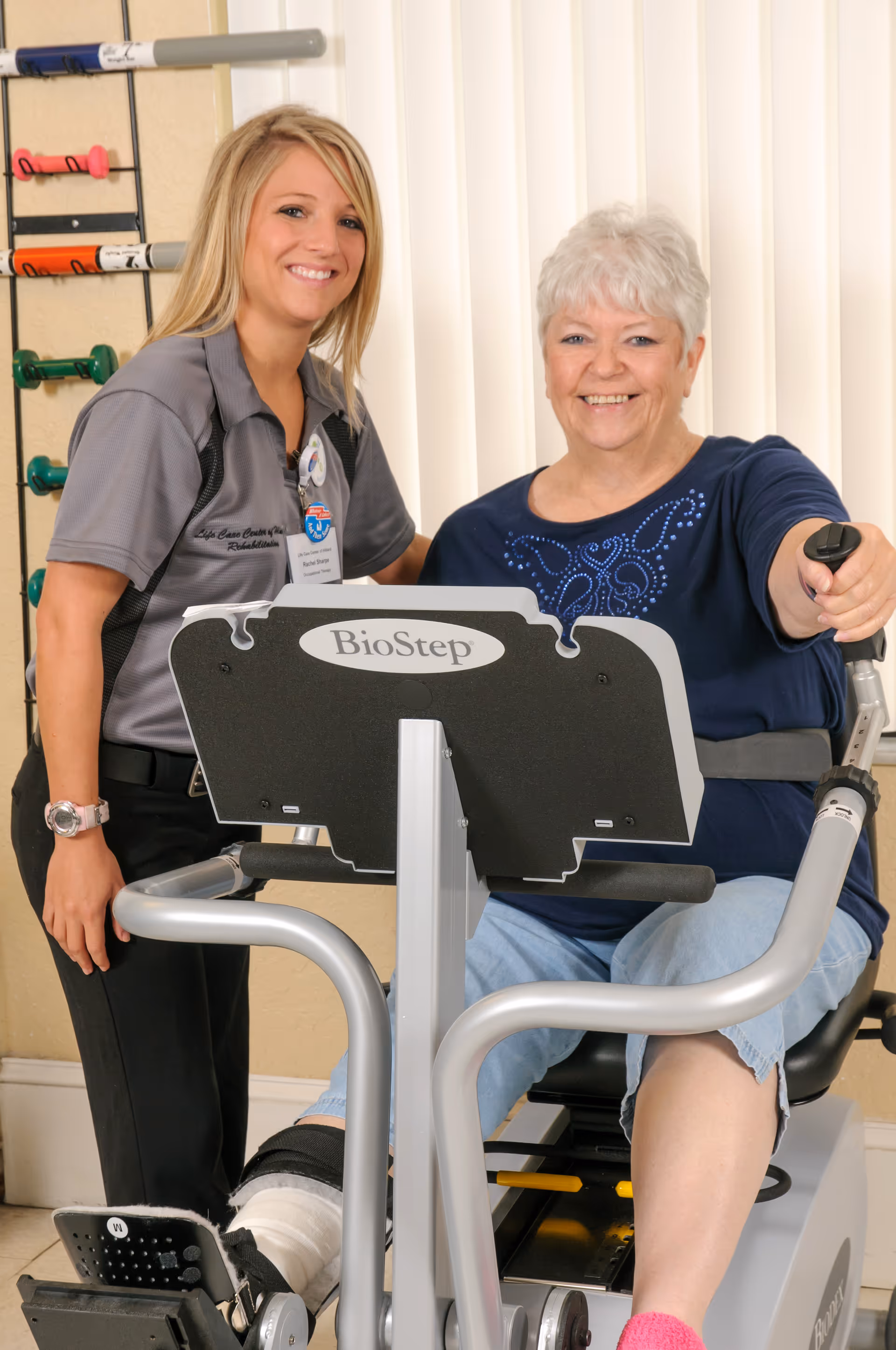A rehabilitation therapist assists an elderly woman using a BioStep exercise machine in a therapy room. The elderly woman is smiling and appears engaged in the exercise, while the therapist stands beside her, also smiling. Various exercise equipment is visible in the background.