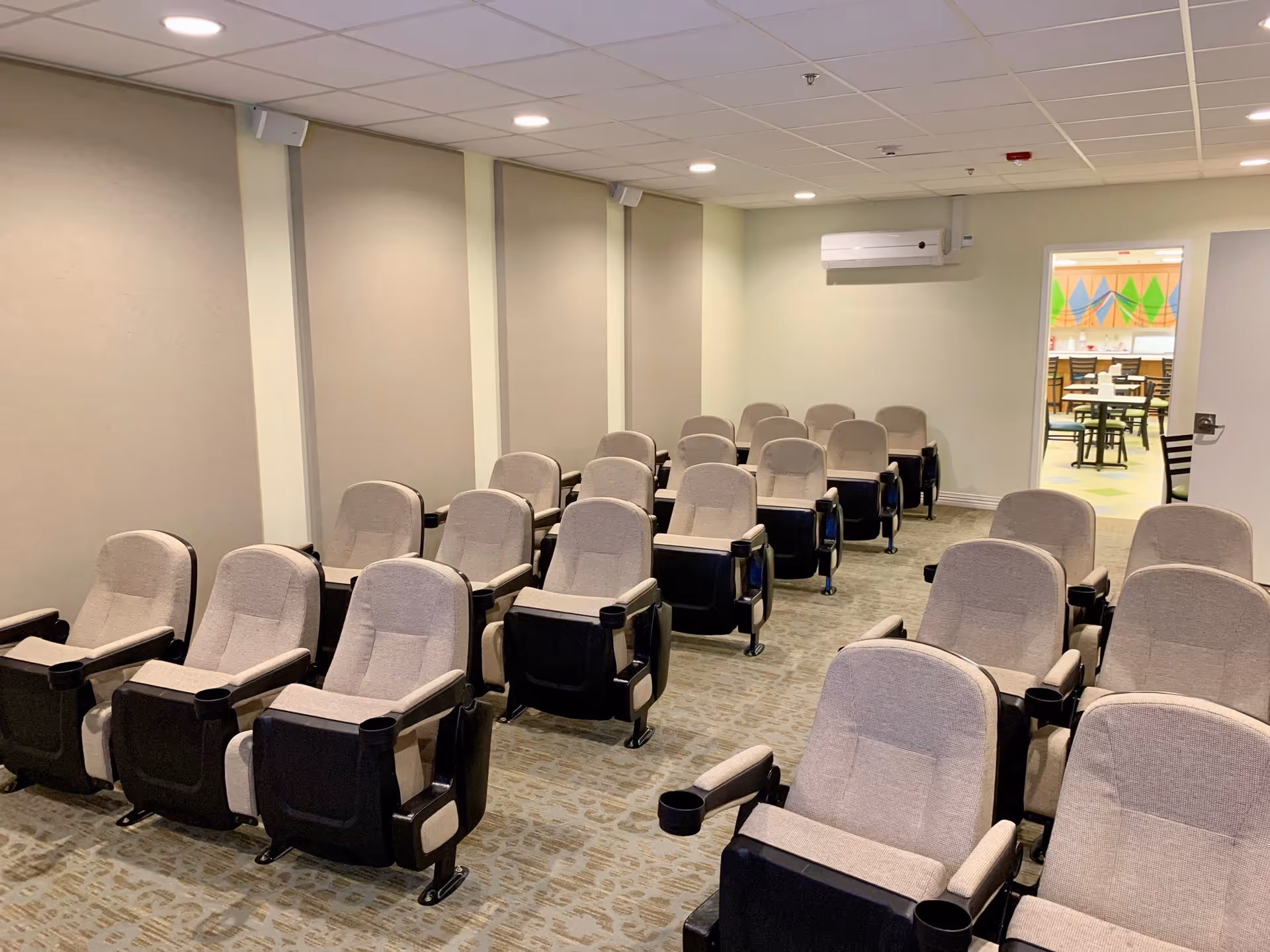 A small theater-style room with three rows of beige cushioned chairs with black armrests and cup holders, arranged facing the front. The room has beige walls with soundproof panels and a carpeted floor. In the background, an open door reveals a brightly colored room with tables and chairs.