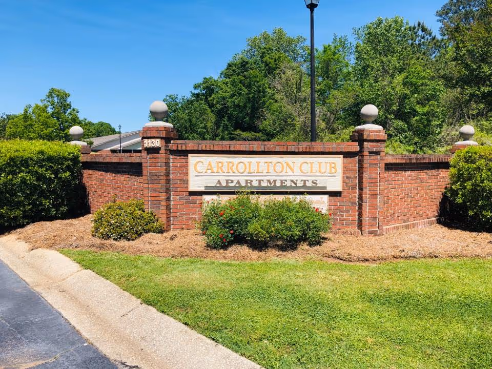 Brick entrance sign for Carrollton Club Apartments surrounded by green bushes and trees under a clear blue sky.
