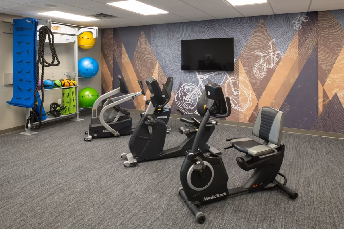 Indoor fitness room with exercise equipment including recumbent stationary bikes and a treadmill. The room has a patterned wall with bicycle illustrations and a mounted flat-screen TV. Various exercise balls and fitness accessories are stored on racks in the corner.