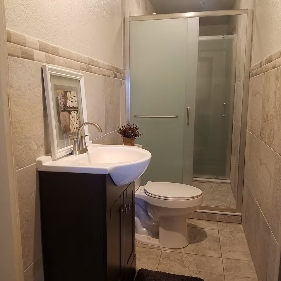 A small bathroom with beige tiled walls and floor. It features a white sink with a dark brown cabinet, a framed picture resting on the sink, a small potted plant, a white toilet, and a shower with frosted glass sliding doors.