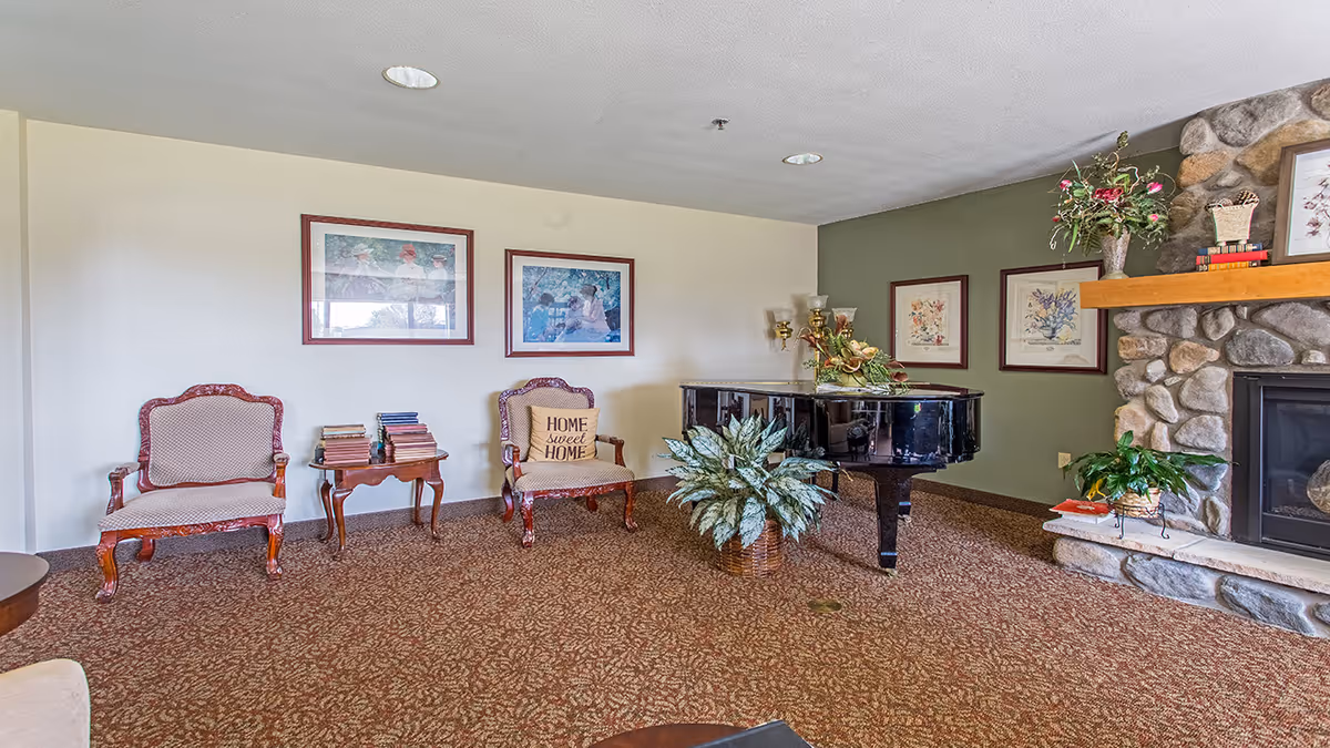 A cozy living room area with two upholstered armchairs, one with a 'Home Sweet Home' pillow, a small wooden table stacked with books, a black grand piano with a floral arrangement on top, a large potted plant in front of the piano, and a stone fireplace with a wooden mantel decorated with flowers and framed artwork on the walls.