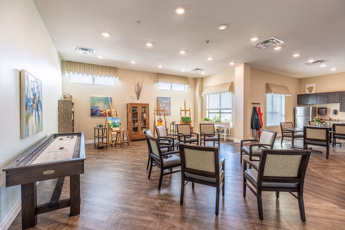 Bright communal activity room with chairs arranged in a circle, an art easel area, a shuffleboard table, and a kitchenette in the background.