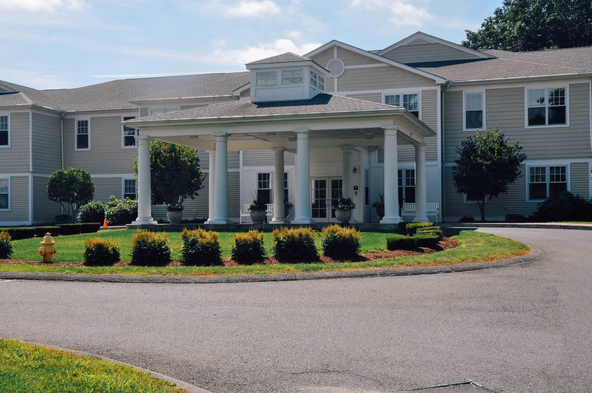 Front entrance of a beige multi-story assisted living building with a covered portico supported by columns, circular driveway, and landscaped shrubs.