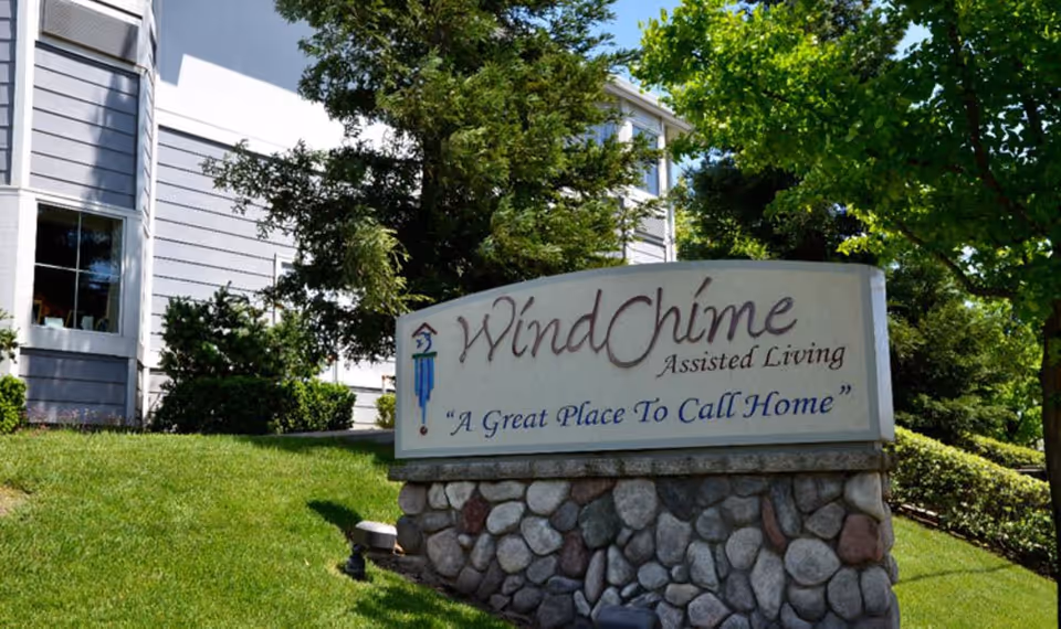 Stone-based sign for WindChime Assisted Living reading "A Great Place To Call Home" on a landscaped lawn with the facility building and trees in the background.