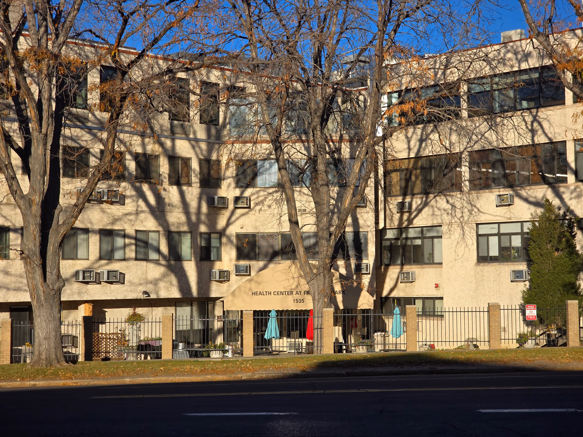 Exterior view of the Health Center at Franklin Park building with multiple windows and air conditioning units. There are leafless trees casting shadows on the building, a fenced patio area with umbrellas, and a clear blue sky.