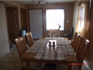 A dining room with a wooden table covered by a beige and brown checkered tablecloth. Six wooden chairs surround the table. A window with curtains is on the right side, and a door and another window are visible in the background. The room has beige walls and a ceiling light fixture.