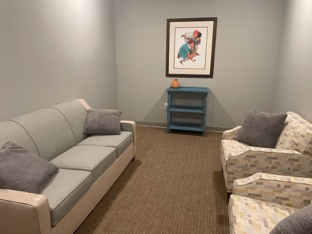 A small sitting room with a light gray couch on the left and two patterned armchairs on the right, each with gray cushions. At the far end of the room is a small blue shelf with a decorative item on top, and a framed picture of two children playing is hung on the wall above the shelf. The walls are painted light gray and the floor is carpeted in a brown tone.