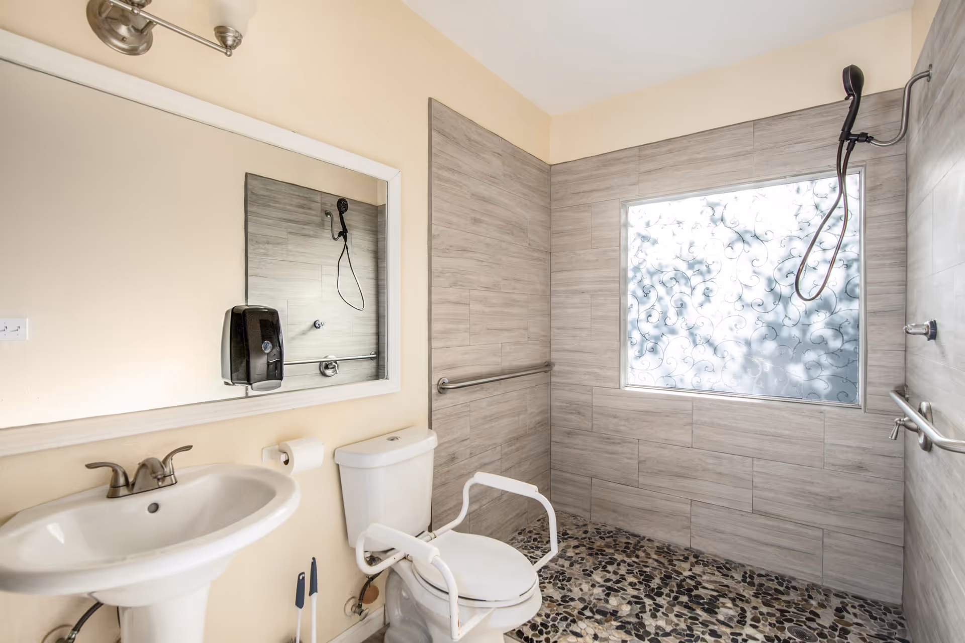 A bathroom with a pedestal sink, a toilet equipped with white safety rails, a large mirror above the sink, and a walk-in shower area with a handheld showerhead and textured glass window. The walls are tiled in a light gray color and the floor has a pebble stone design. Safety grab bars are installed near the toilet and in the shower area.