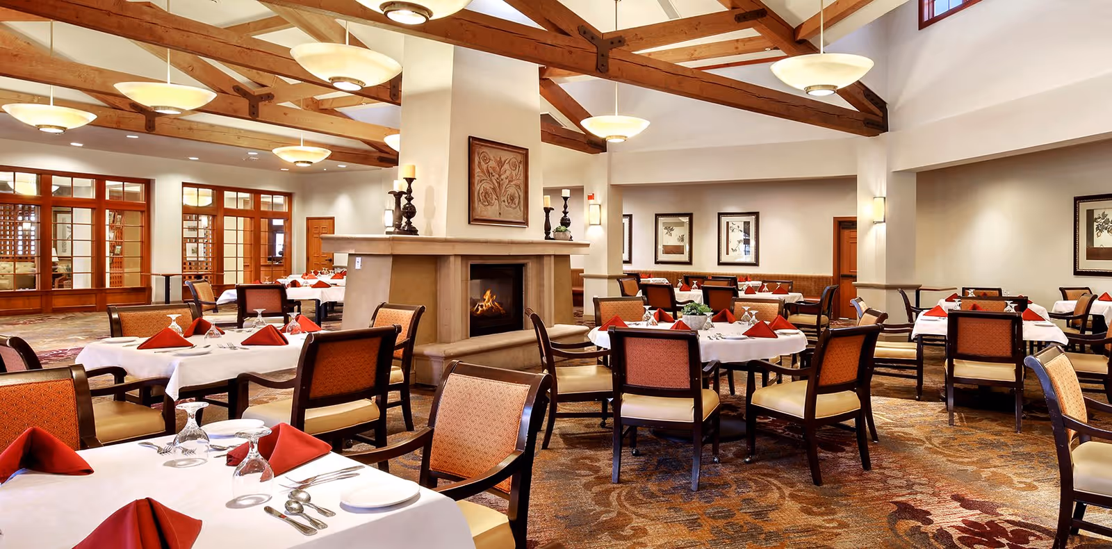 Spacious dining room with multiple tables set with white tablecloths and red napkins around a central fireplace under exposed wooden ceiling beams.