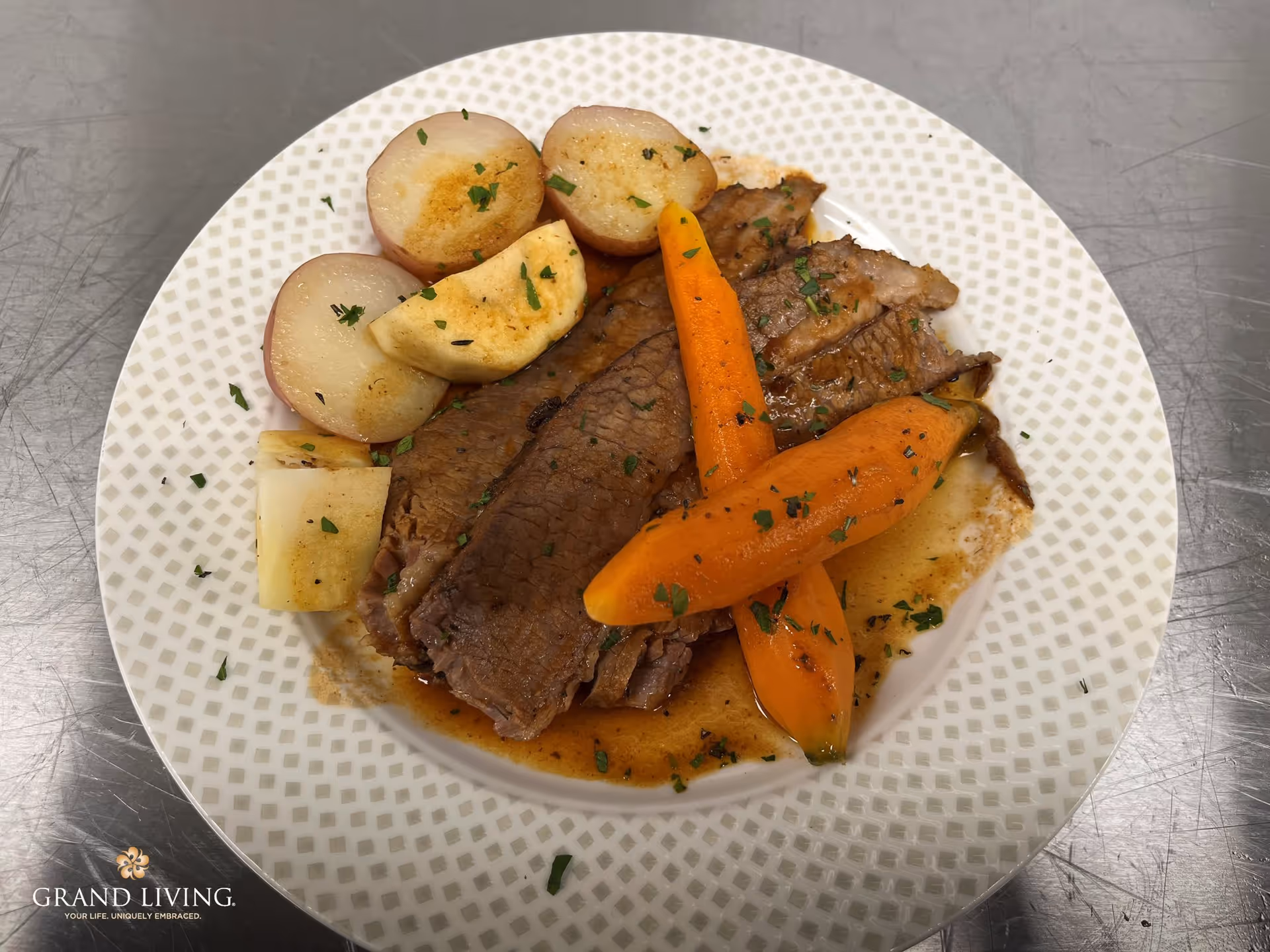 A plate of cooked meal featuring slices of roast beef with gravy, baby carrots, red potatoes, and pieces of turnip garnished with chopped herbs on a patterned white plate.