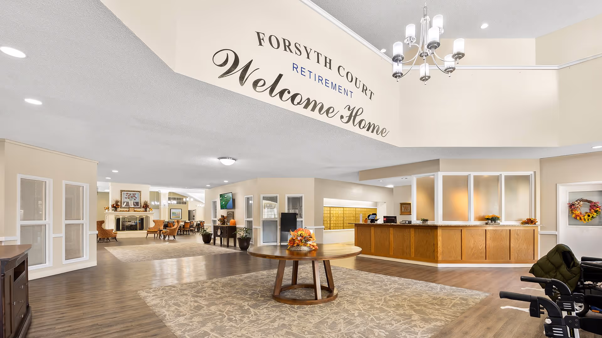 Spacious and well-lit retirement community lobby with a round wooden table decorated with a floral arrangement in the center. To the right is a reception desk with a few small plants and a wreath on the door behind it. The far end features a seating area with chairs and a fireplace. The wall above the lobby entrance has the text 'Forsyth Court Retirement Welcome Home'.