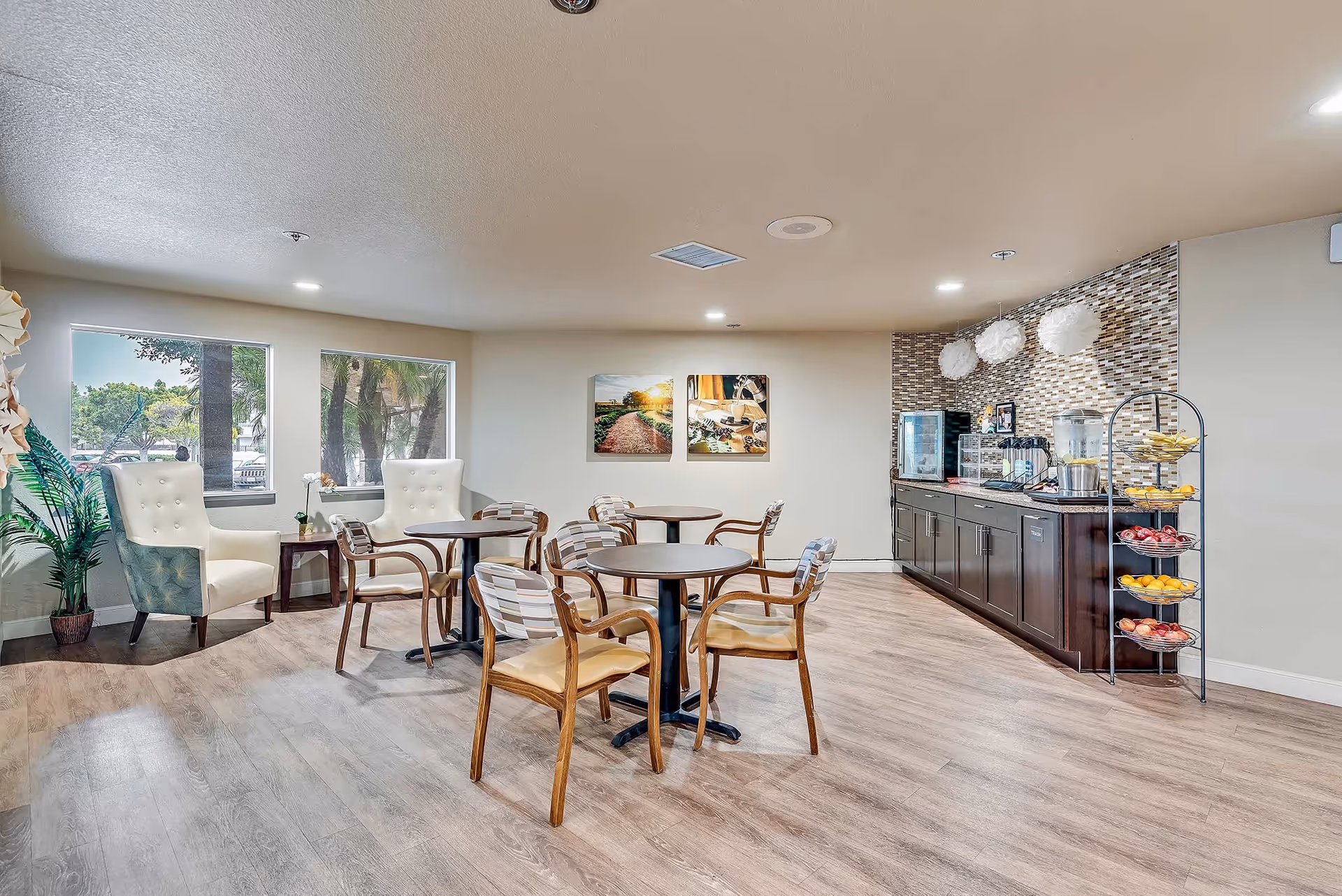 A bright and clean dining area in a senior living facility with several round tables and wooden chairs with cushioned seats. There are two large white armchairs near two windows showing outdoor greenery. On the right side, there is a counter with a mosaic tile backsplash, a beverage dispenser, a small refrigerator, and a rack filled with fresh fruit. The floor is wood-style laminate, and the walls are light-colored with two framed pictures hanging.