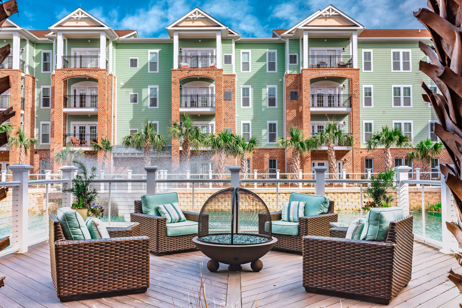 Outdoor seating area with four wicker armchairs with green cushions arranged around a circular fire pit on a wooden deck. In the background, there is a multi-story building with green siding and red brick accents, balconies, and palm trees. A water feature is visible behind the railing surrounding the deck.