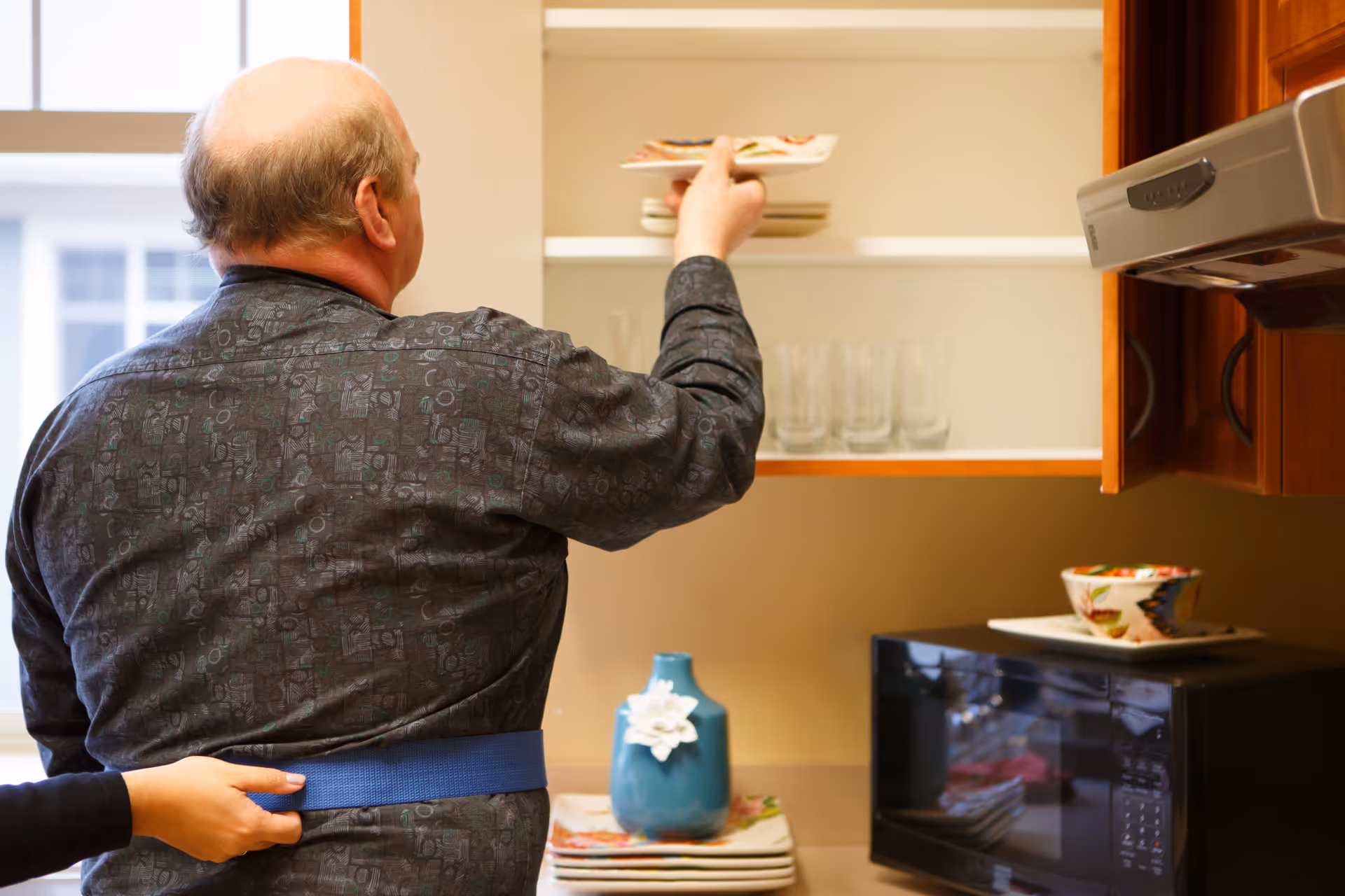 A person reaches into an upper kitchen cabinet while another person steadies them with a blue gait belt.