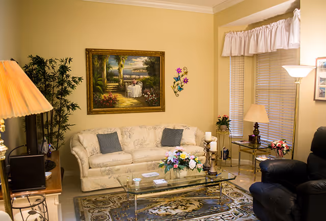 Cozy living room with a floral-patterned sofa, glass coffee table with a flower arrangement, armchair, lamps, and framed wall art.