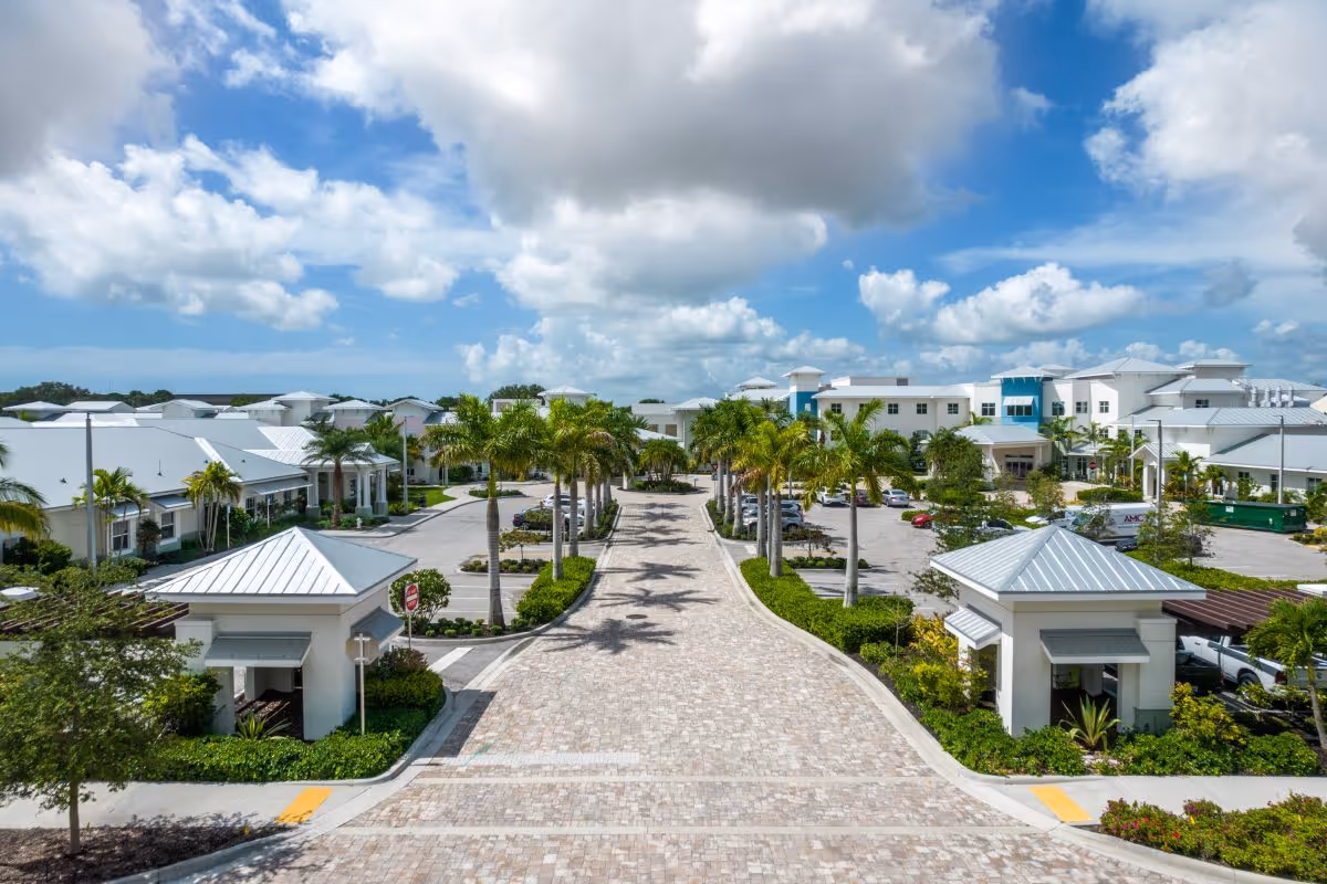 Wide paved driveway lined with palm trees leading to a large, modern senior living facility with white buildings and blue accents under a partly cloudy sky.
