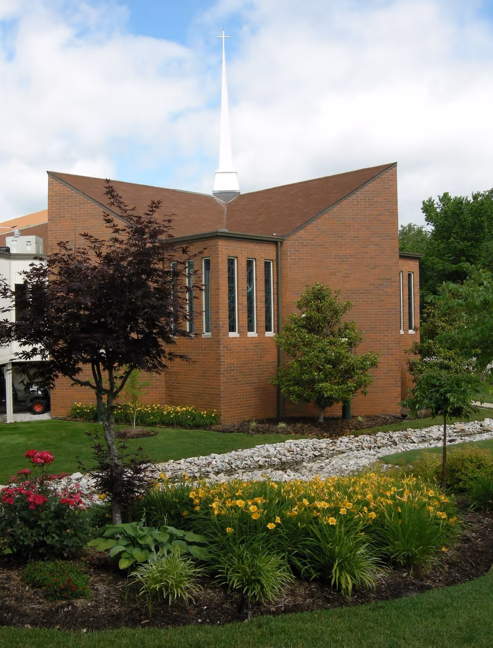 A brick building with a tall white steeple topped with a cross, surrounded by a landscaped garden featuring green grass, yellow flowers, and various trees under a partly cloudy sky.