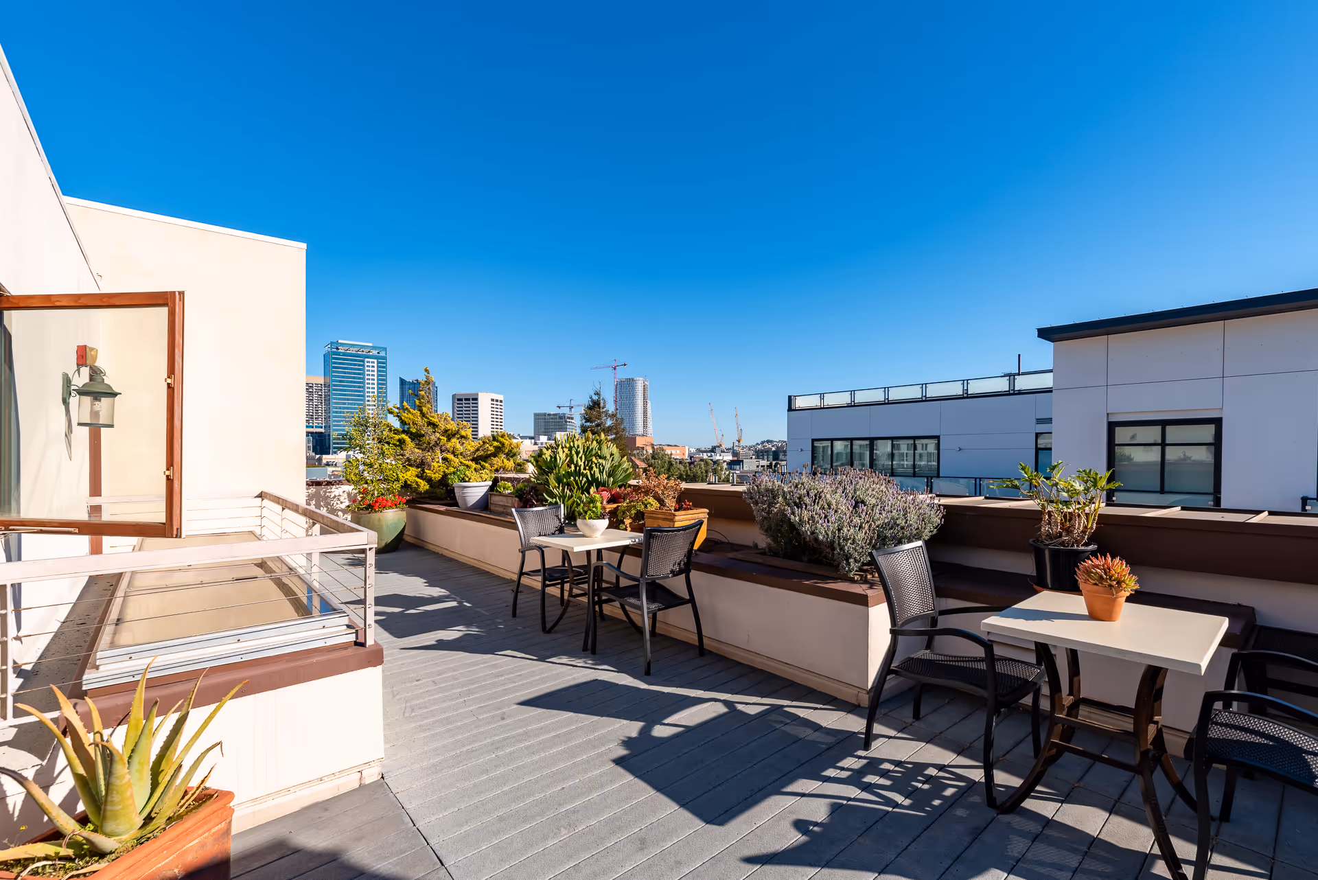 Outdoor rooftop patio area with tables and chairs, potted plants along the railing, and a clear blue sky with city buildings in the background.