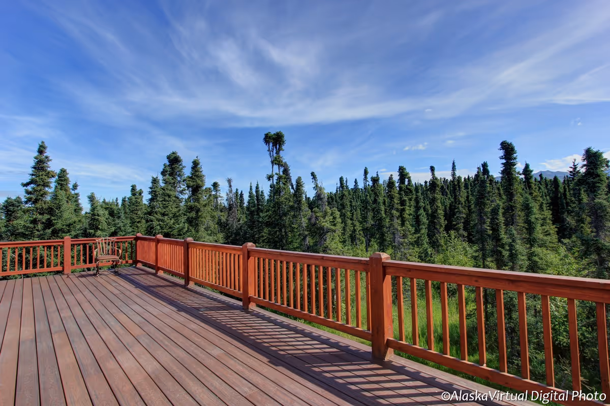 A spacious wooden deck with a railing overlooking a dense forest of tall evergreen trees under a partly cloudy blue sky. There is a single chair placed in the corner of the deck.