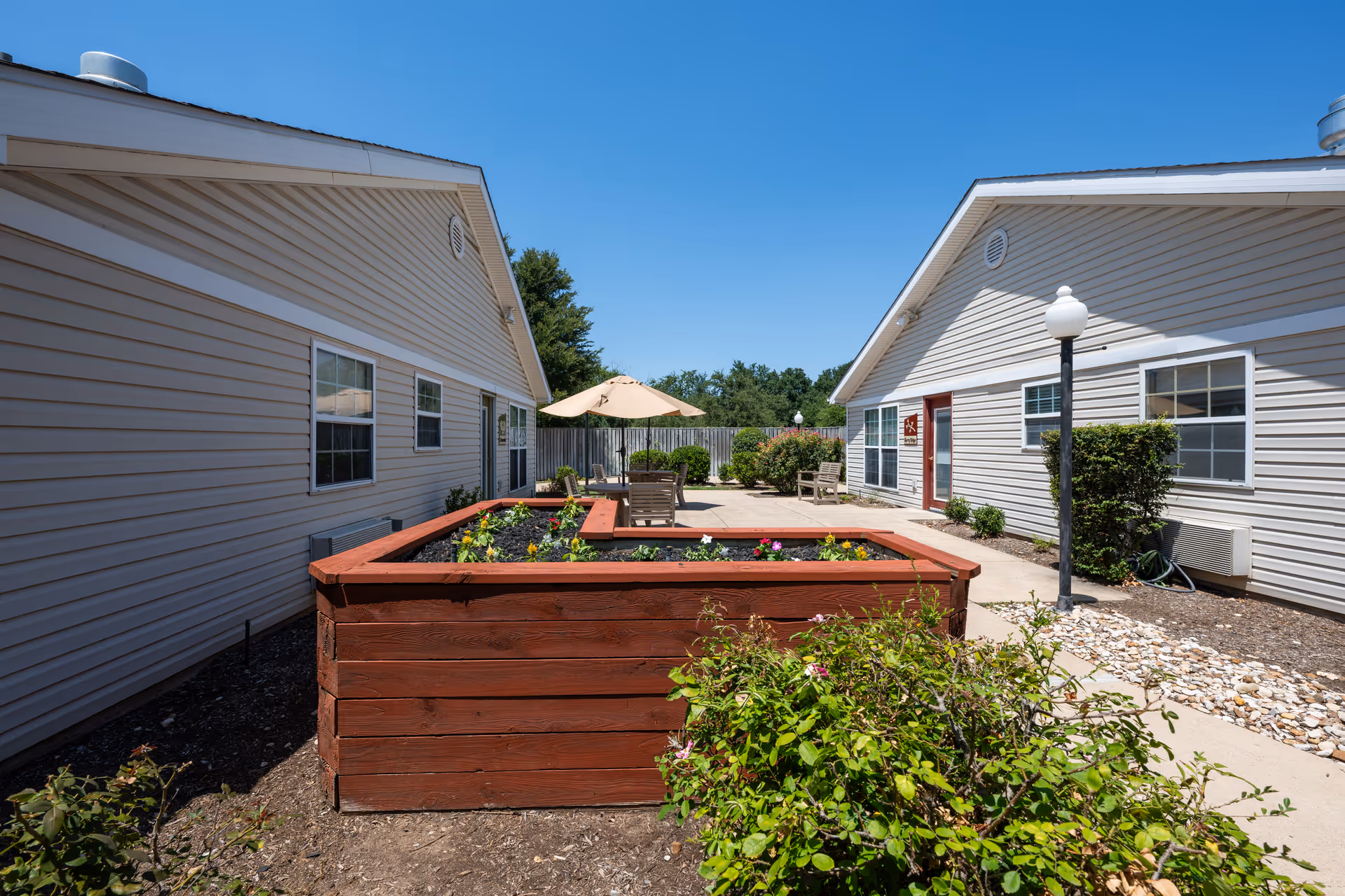 Sunny courtyard between two single-story buildings featuring a large raised wooden planter, patio seating with an umbrella, and landscaping.