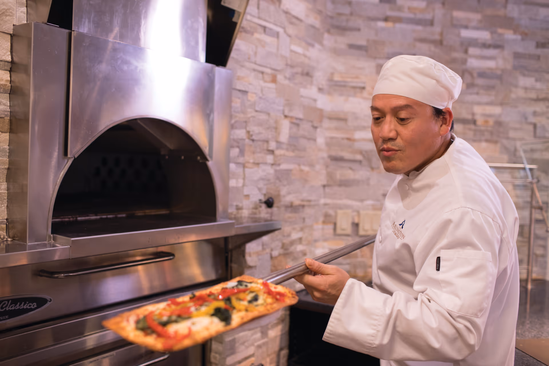 A chef wearing a white uniform and hat is using a pizza peel to place or remove a rectangular pizza with vegetables from a stainless steel pizza oven in a kitchen with stone tile walls.