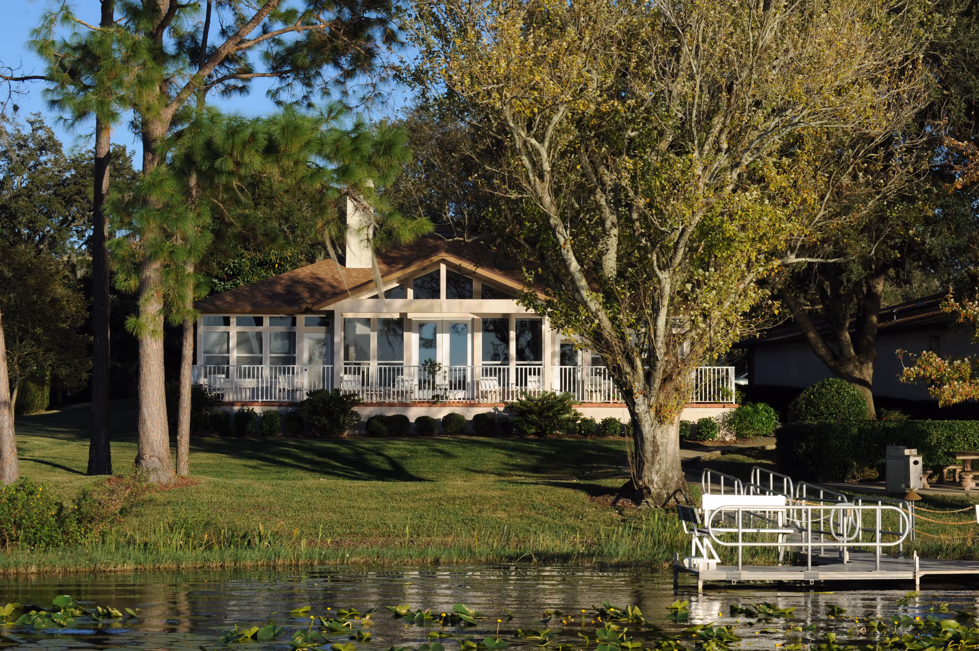 A single-story building with large windows and a porch railing, surrounded by trees and greenery, viewed from across a pond with lily pads and a small dock in the foreground.