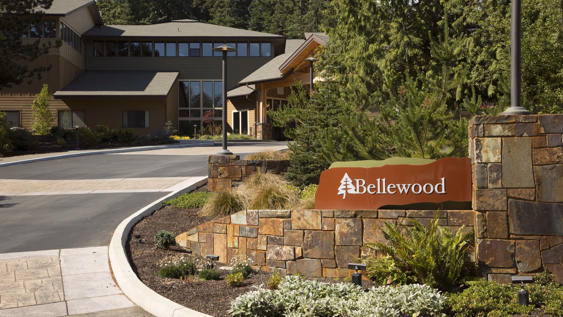 Entrance driveway to Bellewood senior living facility with a stone wall and a sign reading 'Bellewood' surrounded by greenery and trees, with the building visible in the background.
