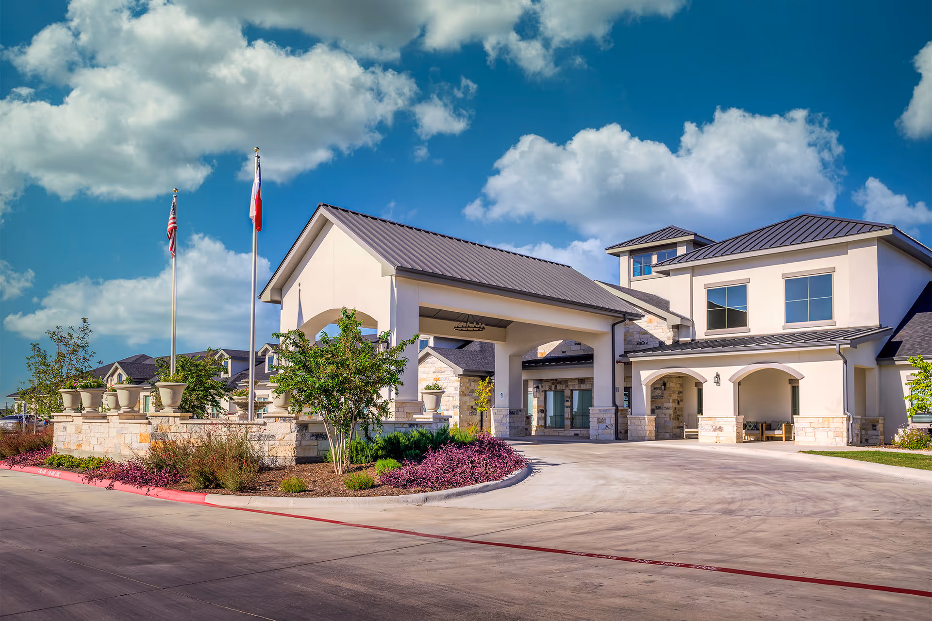 Exterior view of a senior living facility building with a covered entrance, stone and stucco facade, two flagpoles with American and Texas flags, landscaped plants, and a clear blue sky with clouds.