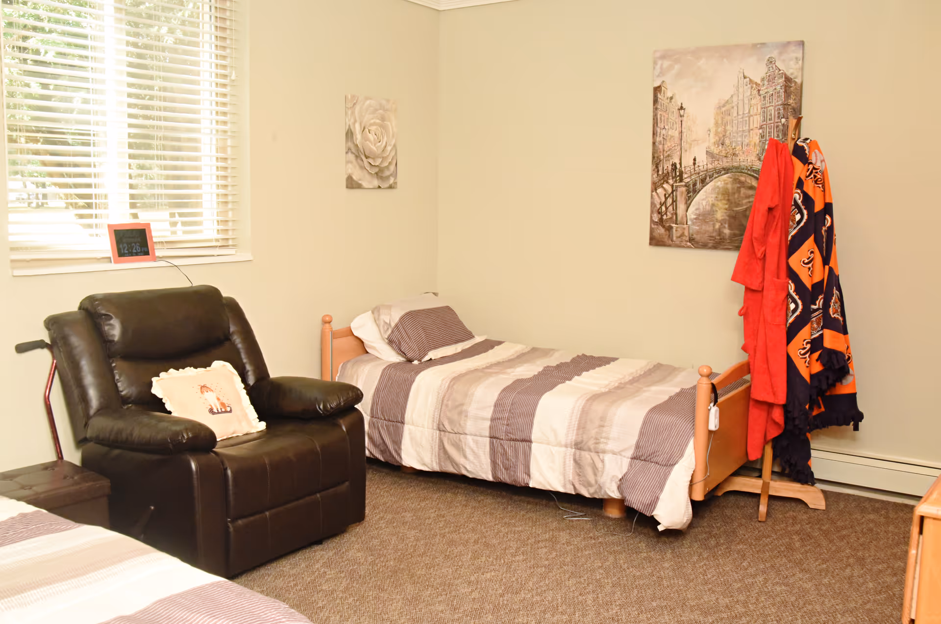 A simple senior bedroom with a single bed, a black recliner, window blinds, wall art, and a coat rack holding blankets and robes.