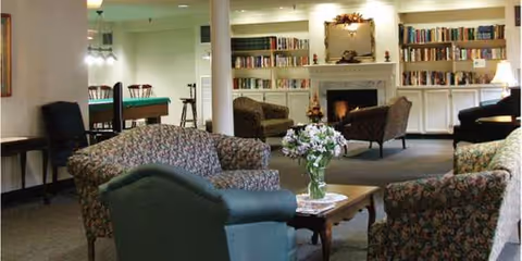 Cozy communal lounge with upholstered chairs and sofas around a coffee table and fireplace, bookshelves and a pool table in the background.