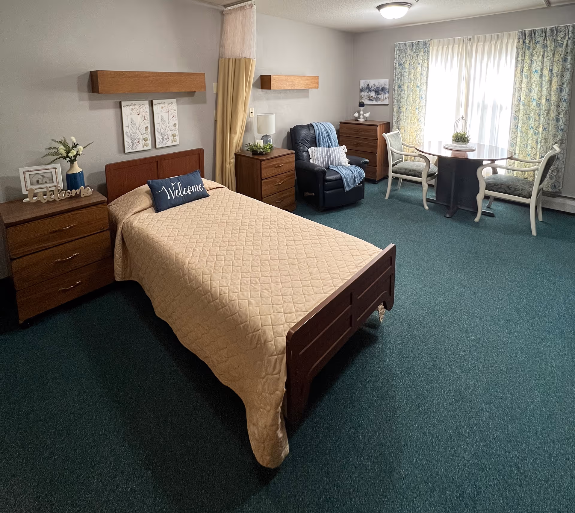 A neatly arranged bedroom in a senior living facility with a single bed covered in a beige quilt and a 'Welcome' pillow. The room features two wooden nightstands with decorative items, a comfortable black recliner chair with a blue throw and pillow, a wooden dresser, and a round table with two chairs near a window with patterned curtains. The carpet is green and the walls are painted light gray.