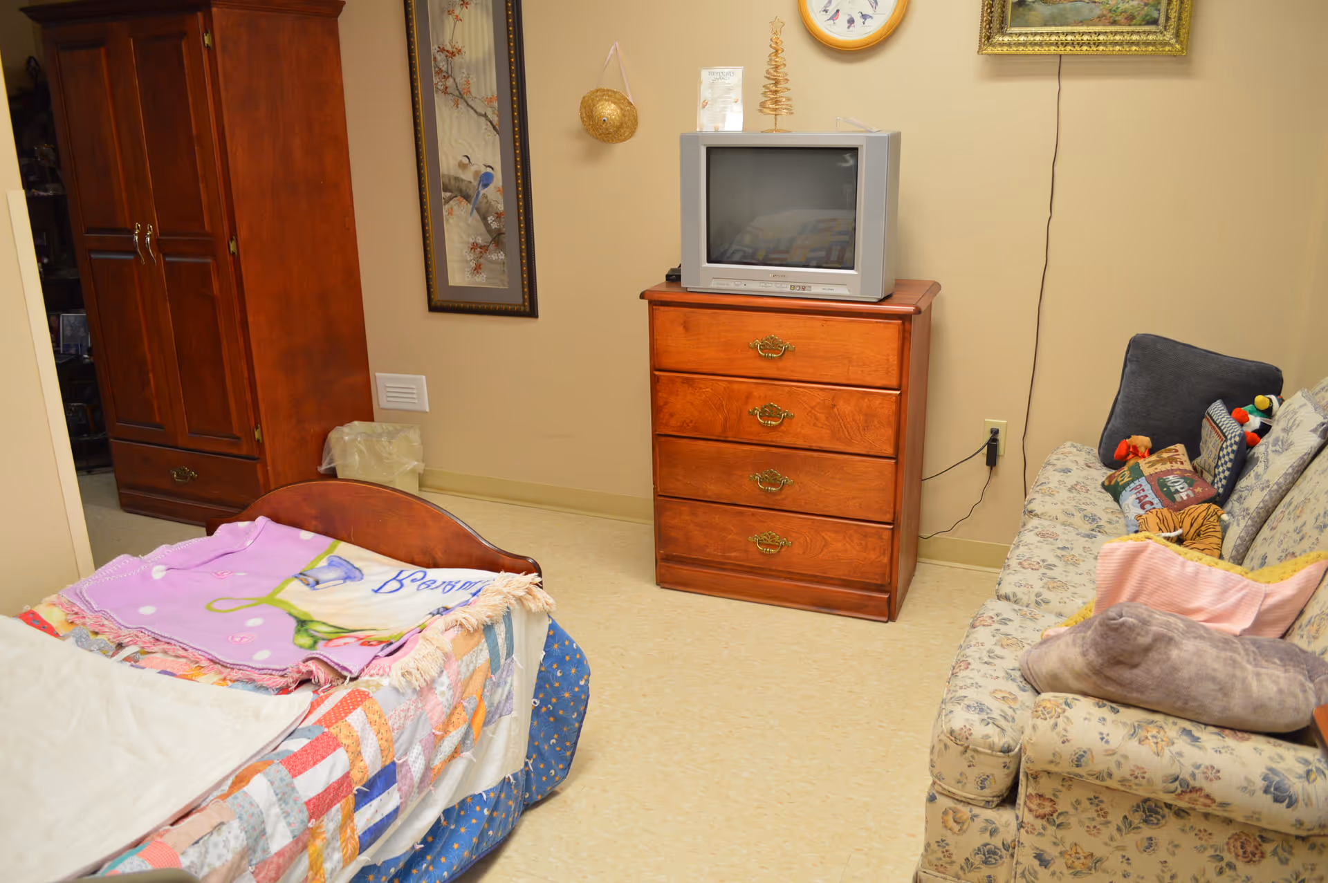 A cozy bedroom in Avoca Lodge featuring a bed with a colorful patchwork quilt and a pink blanket, a wooden dresser with a small CRT television on top, a floral patterned couch with various pillows, and a wooden wardrobe. The walls are decorated with framed artwork and a small hanging basket.
